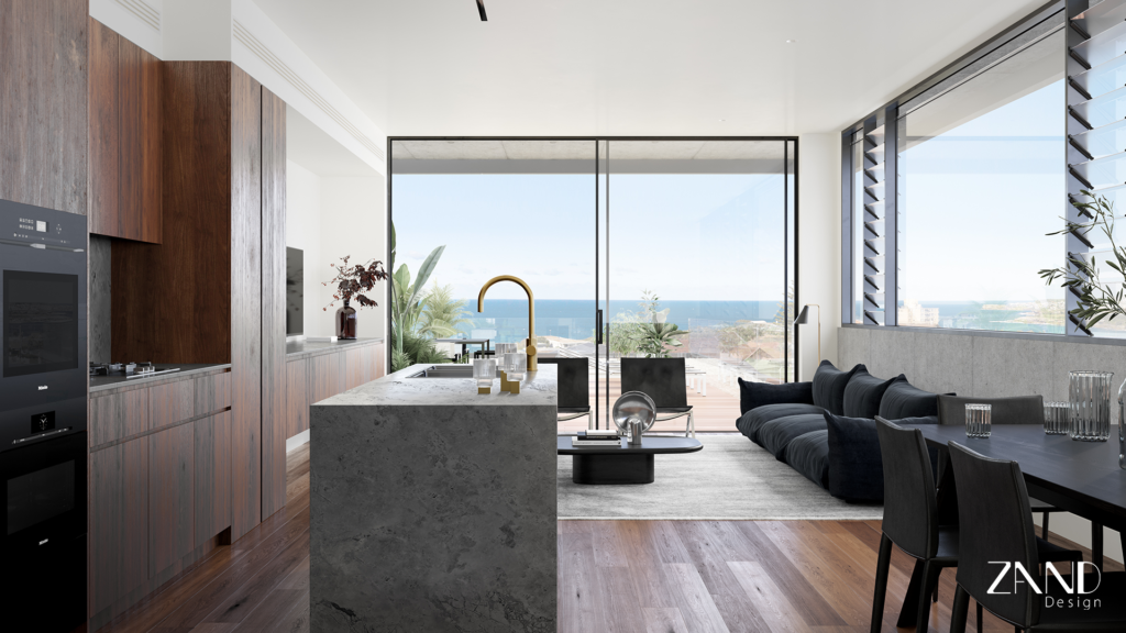 A bright, open-plan interior showing a kitchen with a massive grey stone island and brushed gold tapware. The space flows into a living area with a dark sofa and floor-to-ceiling sliding glass doors that reveal a panoramic view of the coastline and ocean.