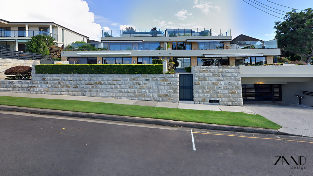 A wide-angle exterior view of a modern, multi-level luxury residence. The building features expansive floor-to-ceiling glass windows and flat roofs with glass balustrades. A prominent, light-colored sandstone block retaining wall runs along the front of the property, topped with a manicured green hedge. In the foreground, there is a paved sidewalk, a strip of green grass, and an asphalt road. A driveway on the right leads to a dark-gated underground garage.