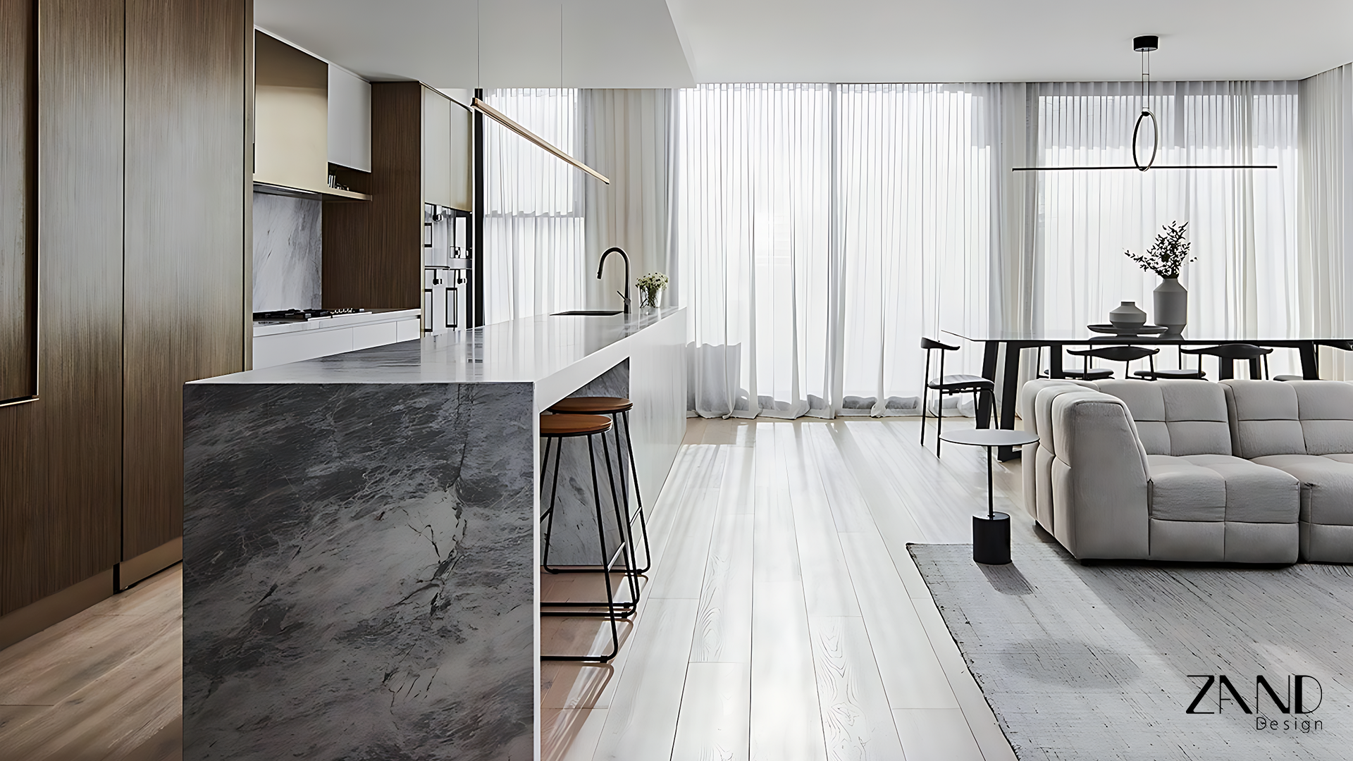A bright, minimalist open-plan interior featuring a kitchen and living area. The focal point is a large marble waterfall island bench with slim black-framed bar stools. To the left, there are sleek wood-paneled cabinets and a marble backsplash. The living area includes a low-profile, grey tufted modular sofa on a textured rug. Floor-to-ceiling sheer white curtains cover large windows, diffusing soft, natural light across the light-toned timber floors. A thin, contemporary linear pendant light hangs over the island.